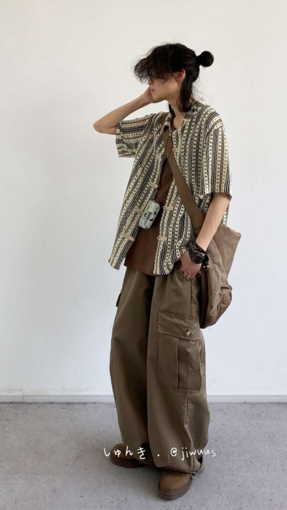 Tousled bun + Tribal-print camp shirt + Rust tee + Brown canvas cross-body + Film-camera holster + Olive cargo wide pants + Brown rugged boots