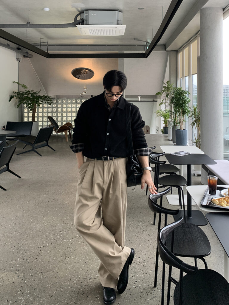Brushed-back fringe + Black knit bomber + Plaid cuff peeks + Coal leather belt + Wide beige pleat trousers + Polished black Chelsea boots