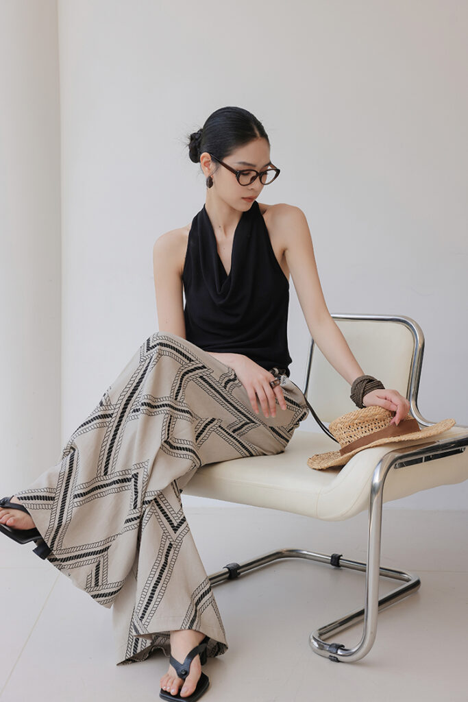 Low bun + tortoiseshell glasses + draped black halter top + beige-and-black geometric flared pants + black flip-flops