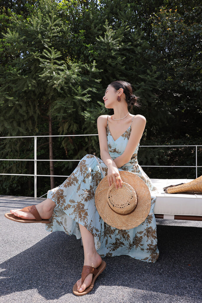 Tousled bun + straw sunhat (carried) + blue floral spaghetti-strap maxi dress + brown leather flip-flops